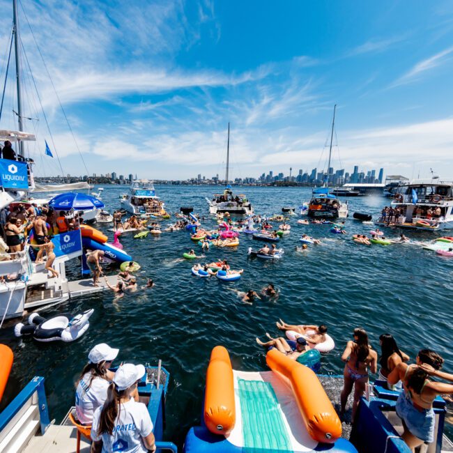 A lively boat party on a sunny day with numerous people in the water on colorful inflatables. Several boats are anchored nearby, and a city skyline and bridge are visible in the background. Many attendees are socializing and enjoying the festive atmosphere.