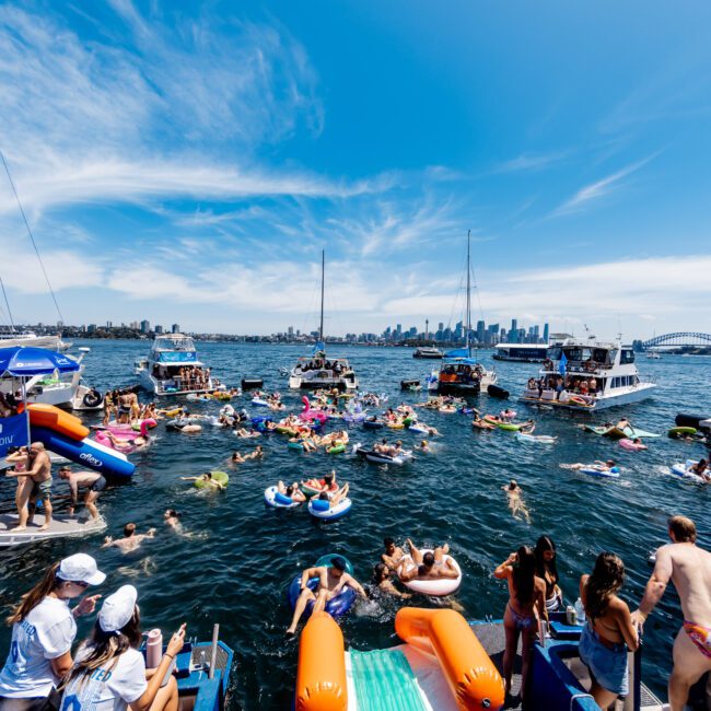 People enjoying a sunny day on a lake filled with inflatable floats and boats. The sky is clear with wispy clouds. The city skyline is visible in the distance. There are groups swimming and socializing on the water and boats.