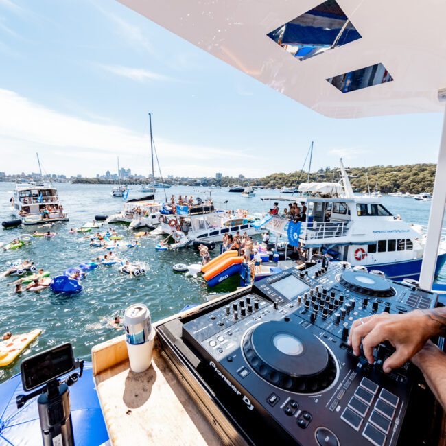 A DJ is playing music on a boat deck during a lively water party. People are swimming and floating on inflatable rafts in the water. Several boats are anchored nearby, and a city skyline is visible in the distance.