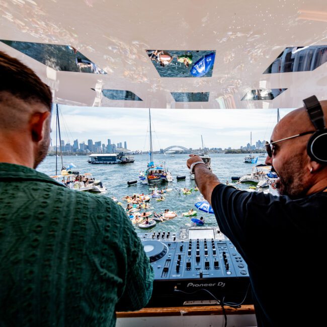 Two DJs perform on a boat overlooking a crowded harbor with numerous boats and inflatables. One DJ wears headphones and points outward. The Sydney skyline, including the iconic bridge, is visible in the background.