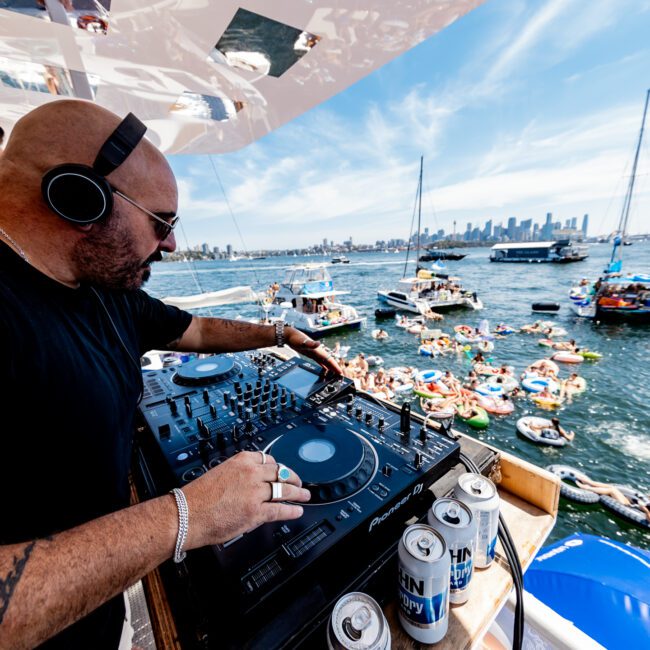 A DJ wearing headphones mixes music on a boat deck, overlooking a lively scene with people on floaties in the water. The background features a cityscape and bridge under a blue sky. Cans of drink are on a table beside the DJ.