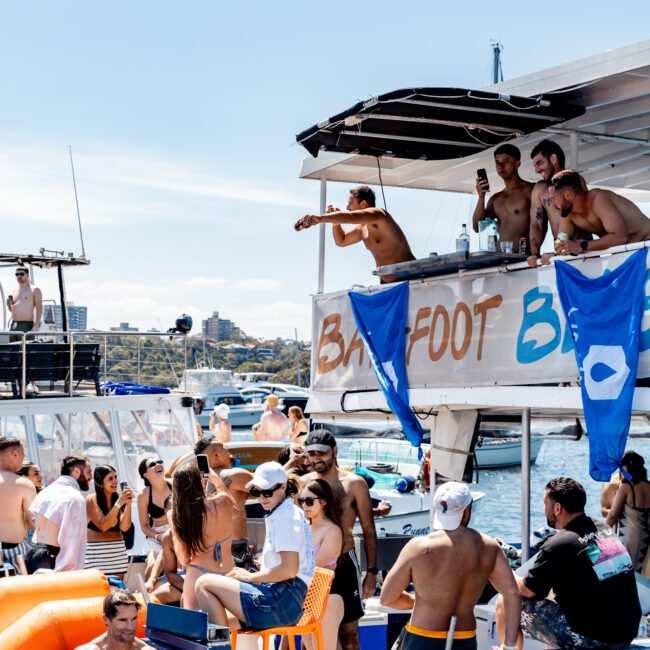 A lively party scene on a boat with people enjoying the sun and socializing. Some are standing, others are seated, and there are inflatable orange and blue decorations around. A shirtless man is seen raising a drink on the upper deck.