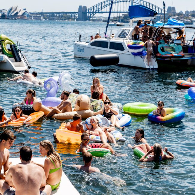 People enjoying a sunny day on Sydney Harbour, floating on inflatables and swimming near anchored boats. The Sydney Harbour Bridge and Opera House are visible in the background, along with a catamaran. The scene is lively and festive.