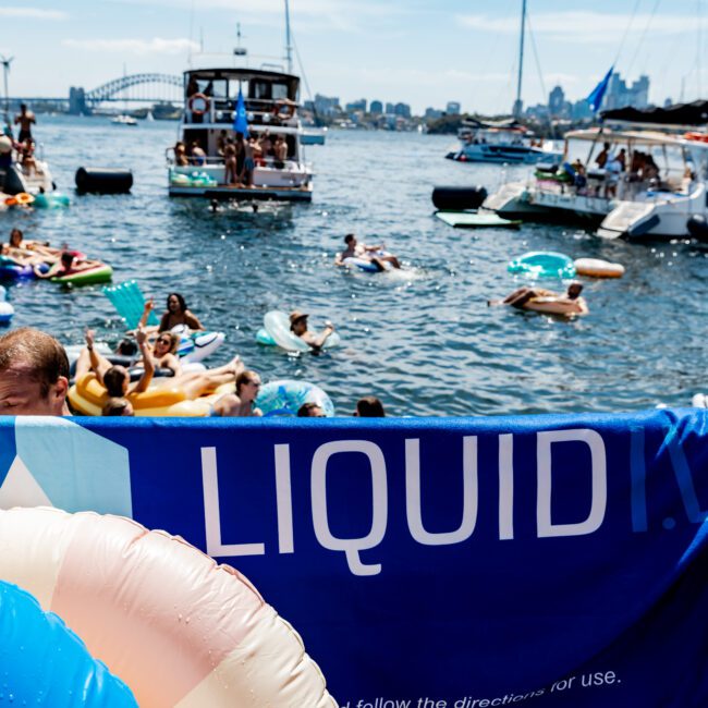 People are lounging on inflatable floats in a sunny harbor surrounded by boats. A banner with the text "LIQUID" is visible in the foreground. In the background, a bridge and skyline are visible under a clear sky.