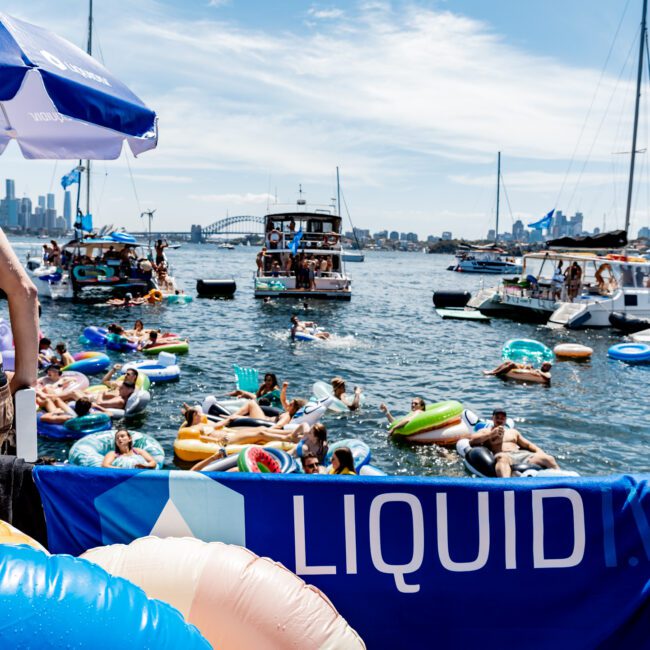 People enjoy a sunny day on a bustling bay filled with colorful inflatables and boats. The city skyline is visible in the background. A blue banner with "LIQUID" is in the foreground, and a large bridge can be seen across the water.