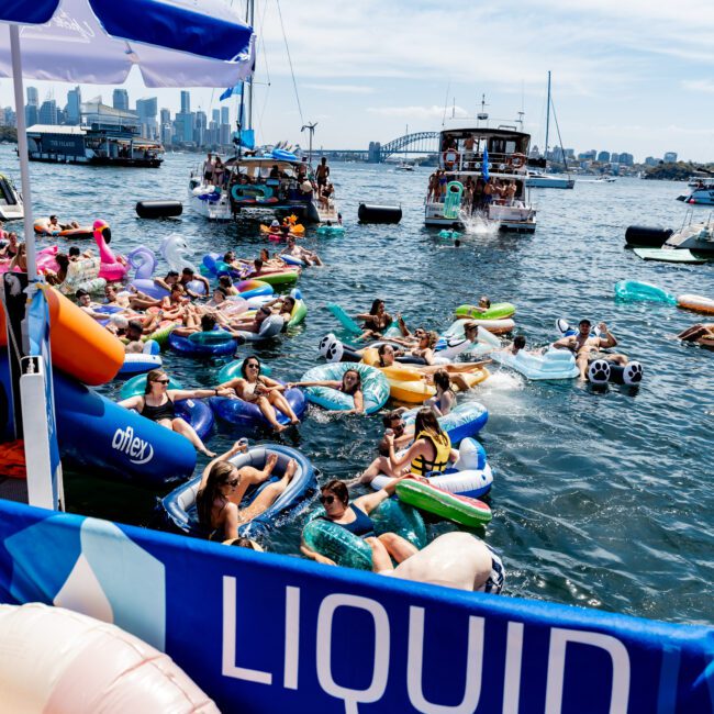 A lively scene of people on colorful inflatable floats in the water, surrounded by boats. An umbrella and banner with "LIQUID" are visible. The background shows a city skyline and bridge under a clear sky, suggesting a festive summer atmosphere.