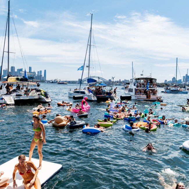 A lively scene of people enjoying a sunny day on the water. Several individuals are seen on inflatable floats and a floating mat near boats. The backdrop features a skyline with skyscrapers under a partly cloudy sky.