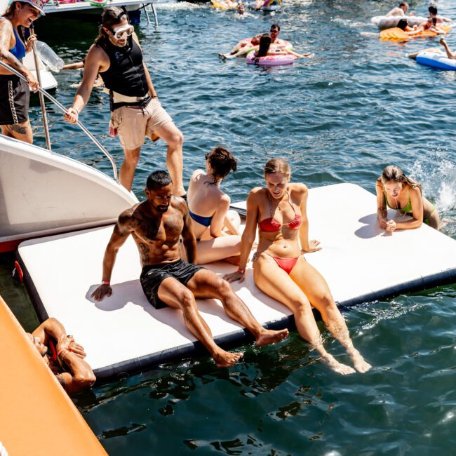 People enjoying a sunny day on a floating platform in the water. Some are sitting, others are swimming or on inflatables. Boats are nearby, and the atmosphere is lively with clear blue skies.