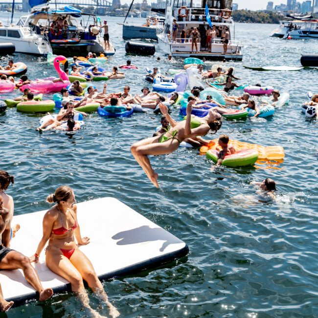 People are enjoying a sunny day on the water. One person is mid-flip into the lake from a floating platform. Others are relaxing on colorful inflatables, with sailboats and a city skyline in the background.