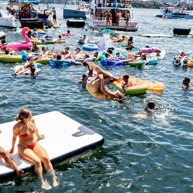 A lively scene on a sunny day with people enjoying the water by floating on inflatable pool toys and platforms. Boats are anchored nearby, and a distant city skyline is visible. A person is flipping into the water from an inflatable.