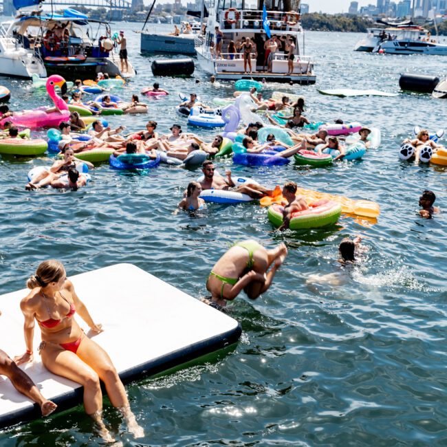 People enjoy a sunny day on the water, with many on colorful inflatables. A person in a green swimsuit dives into the water from a floating platform. Boats are anchored nearby, and a city skyline is visible in the distance.