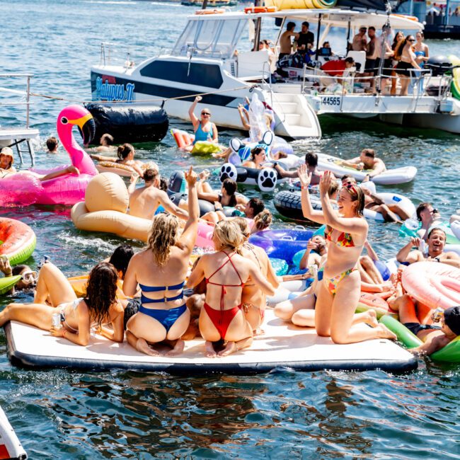 A group of people in colorful swimwear enjoy a summer day on inflatable floats and a boat in a harbor with a city skyline in the background. The scene is lively, with laughter and clear water under a sunny sky.