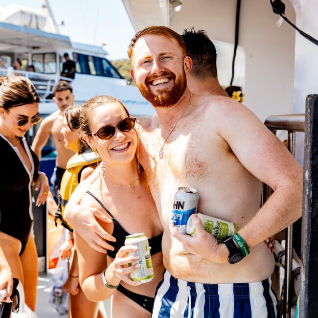 A smiling man and woman in swimsuits stand together on a boat. The man holds a can and wears striped shorts. Other people are in the background enjoying the sunny day, with another boat visible nearby.