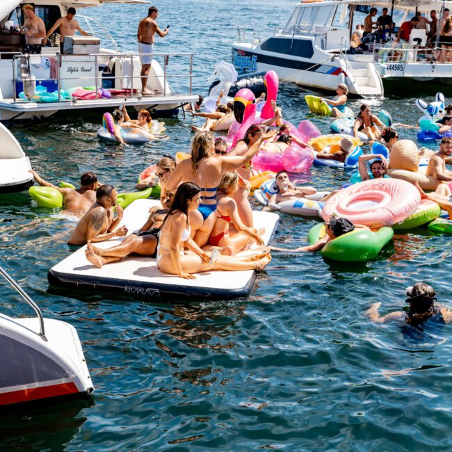 People enjoying a sunny day on a lake with inflatables and boats. They relax on colorful floaties, with some sitting on a large white inflatable platform. The city skyline is visible in the background, and several yachts are docked nearby.