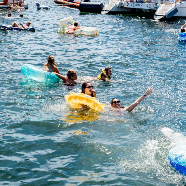 People enjoying a sunny day on the water, swimming and relaxing on inflatable tubes near boats. The scene is lively with several boats in the background and individuals chatting and having fun in the water.