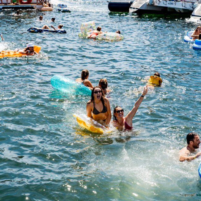 People enjoying a sunny day on a lake, with some on inflatable floats and others swimming. Boats are anchored nearby, and the scene is lively with socializing and relaxation.