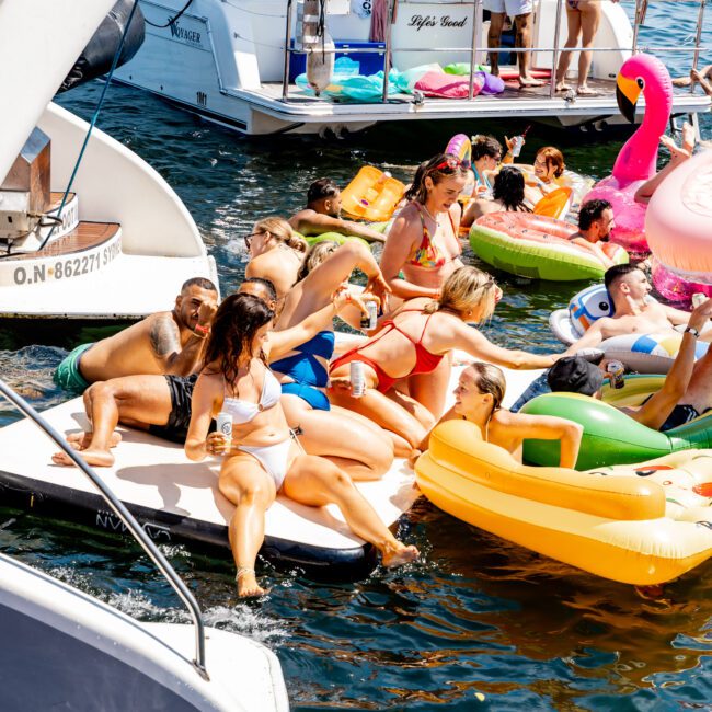 People are enjoying a sunny day on the water, with some lounging on inflatables like a pink flamingo and yellow duck. Others are on boats, socializing and relaxing. The atmosphere is lively and festive.