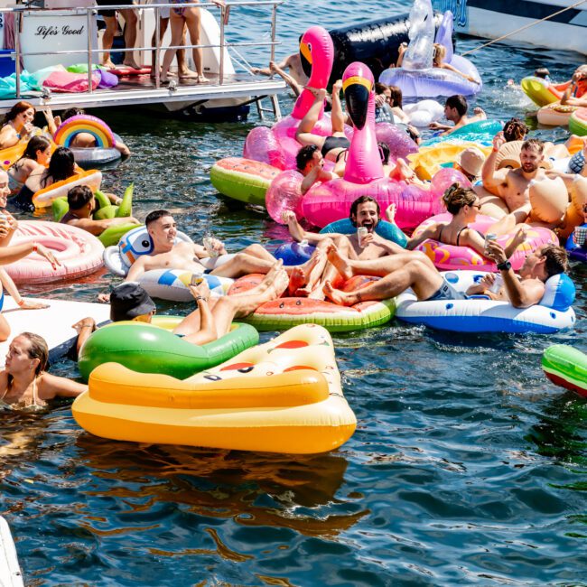 People enjoying a sunny day on colorful inflatable pool floats, including flamingos and unicorns, in the water near boats. The scene is lively and festive, with individuals relaxing and socializing.