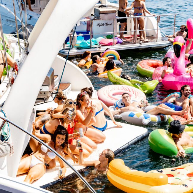 People enjoy a sunny day on boats and colorful inflatables in the water, surrounded by floating unicorns, flamingos, pizzas, and more. Some are relaxing on the deck, socializing, and sipping drinks, creating a lively and festive atmosphere.