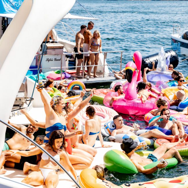 A large group of people enjoying a sunny day on a yacht and in the water, surrounded by colorful inflatable pool floats, including flamingos, rainbows, and unicorns. The atmosphere is lively and festive, with boats visible in the background.