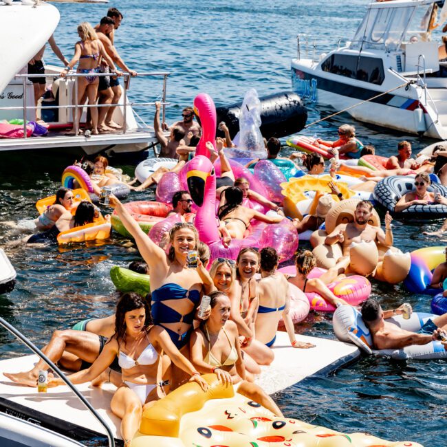 A large group of people enjoying a sunny day on the water, surrounded by a variety of colorful inflatables. They are gathered near boats, with some sitting and others standing on floating platforms. The atmosphere is lively and festive.