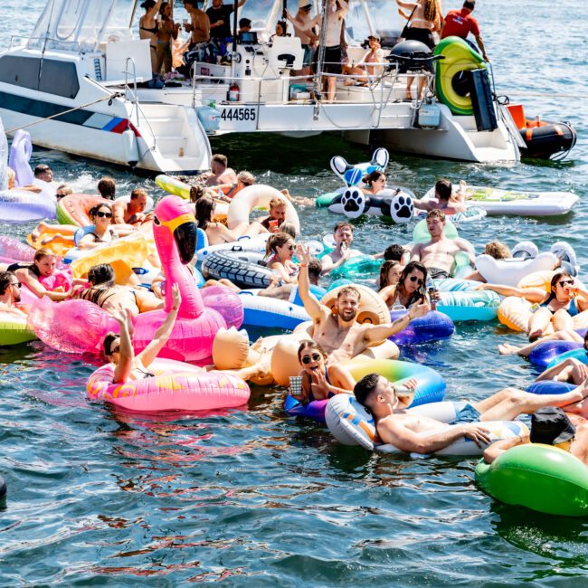 A group of people enjoying a sunny day on colorful inflatable floats, including a flamingo, unicorn, and pizza, near a boat. The city skyline is visible in the background.