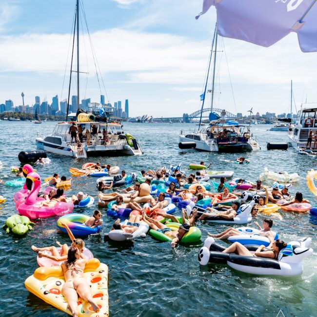 A crowded group of people relax on various inflatable floats, including pizza and flamingo shapes, in a harbor. Sailboats are anchored nearby, and a city skyline and bridge are visible in the background under a sunny sky.