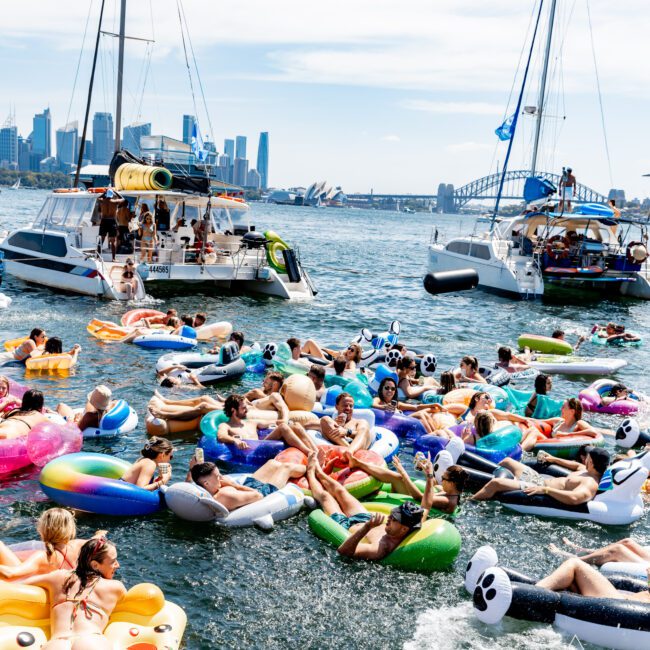 People relaxing on colorful inflatables in the water, surrounded by boats. The background features a city skyline and a prominent bridge. It's a sunny day with clear skies, creating a lively and festive atmosphere.