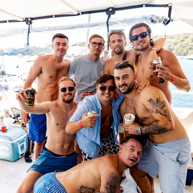 A group of eight shirtless men poses and smiles aboard a boat. Some hold drinks, and they are surrounded by coolers. The background shows a body of water and distant greenery under a sunny sky.