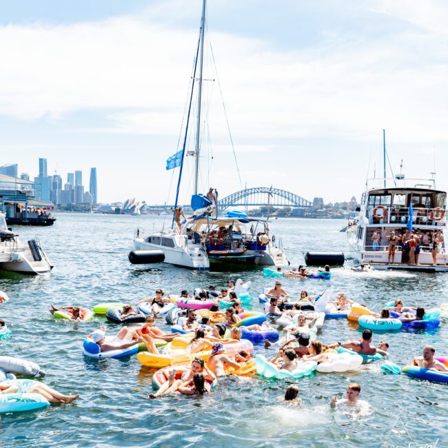 A lively scene shows people in colorful inflatables enjoying the water near anchored boats. The backdrop features a city skyline with the Sydney Harbour Bridge in view. The atmosphere is festive and summery.