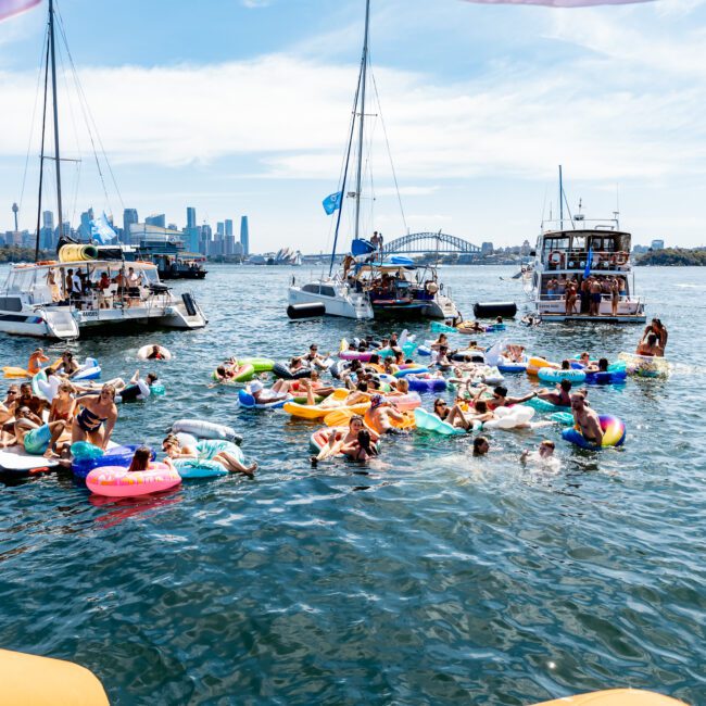 A group of people floating on inflatable toys in a harbor, surrounded by several yachts. The city skyline and a bridge are visible in the background under a clear blue sky.