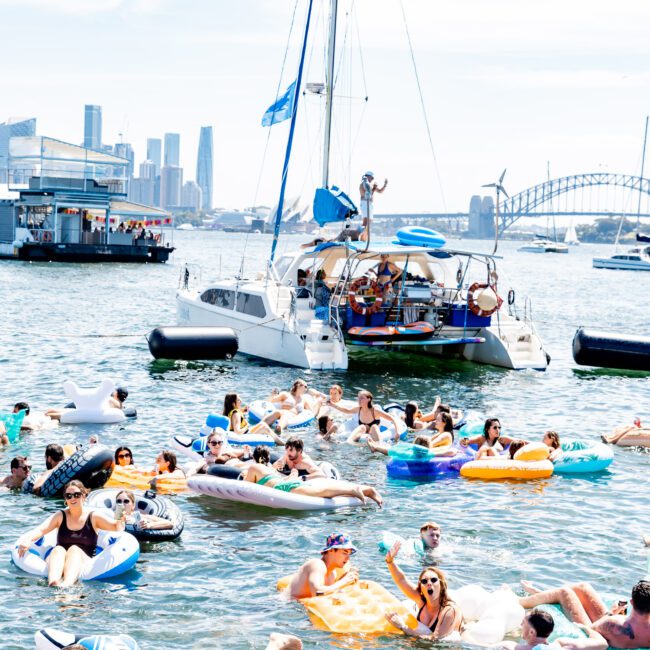 A lively scene on the water with people relaxing on colorful inflatables. A sailboat is in the background, along with a cityscape and a bridge. The weather is sunny and bright, enhancing the festive atmosphere.