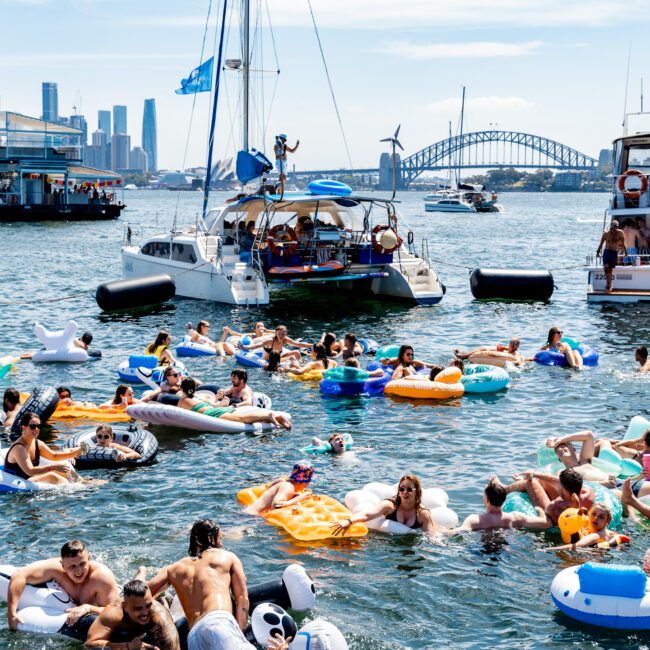 A group of people on colorful floaties enjoy the water near several yachts. The Sydney Harbour Bridge and city skyline are visible in the background under a clear blue sky.