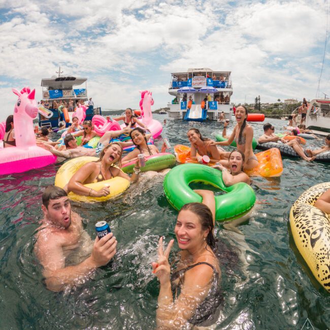 A group of people enjoying a sunny day on the water. They are on inflatable floats, including a pink unicorn, a flamingo, and colorful rings. Some are holding drinks, and there's a boat with more people in the background.