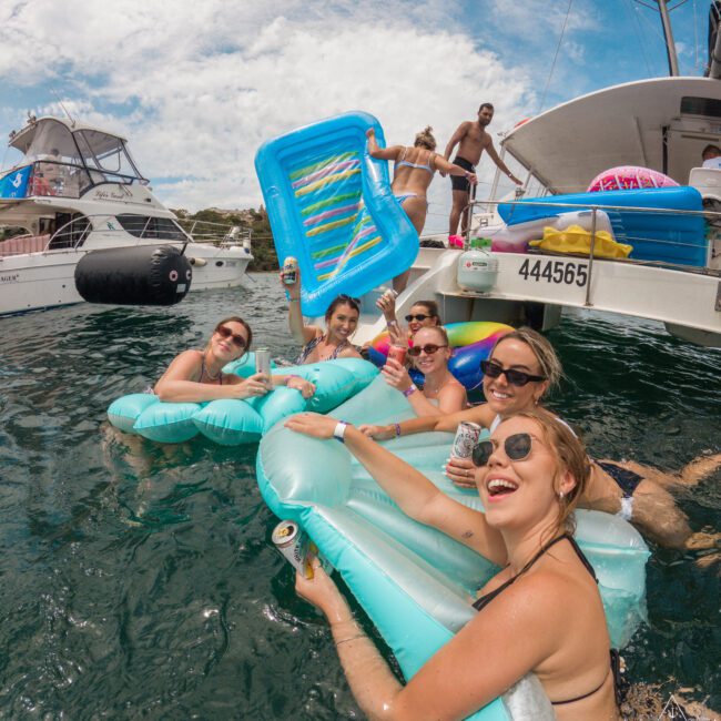 A group of smiling people in swimsuits are relaxing on inflatable floats in the water next to a boat. They are holding drinks and enjoying a sunny day. Other boats are visible in the background under a partly cloudy sky.