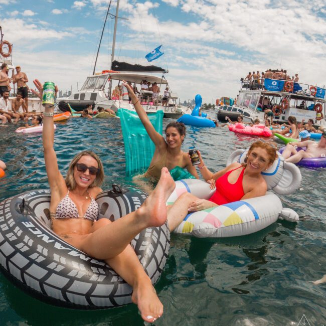 Three women are reclining on inflatable pool floats in the ocean, raising drinks and smiling. Behind them, there are boats and a group of people enjoying the water under a partly cloudy sky.