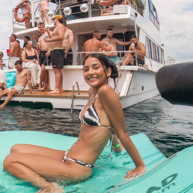 A woman in a striped bikini smiles while sitting on a floating mat in water. In the background, a group of people gather on a boat, some sitting and others standing. The scene is lively and festive.