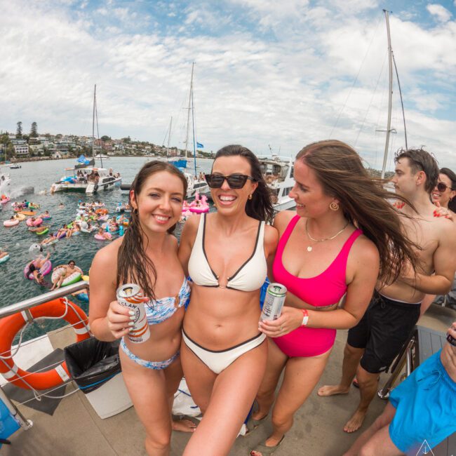 Three women in swimsuits smile and hold drinks on a boat deck with water and other boats in the background. The scene is lively, with people on inflatables and a partly cloudy sky overhead.