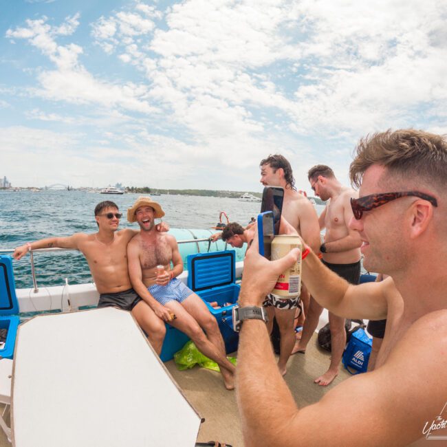 A group of people enjoying a sunny day on a boat. One person holds a drink in the foreground, while others relax and chat on deck. The ocean and a distant city skyline are visible under a partly cloudy sky.