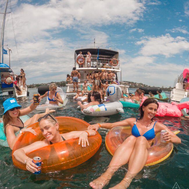 People in swimsuits enjoying a sunny day on a lake. They are floating on inflatables and holding drinks. In the background, others are on boats with more inflatables. The sky is partly cloudy, creating a festive summer atmosphere.