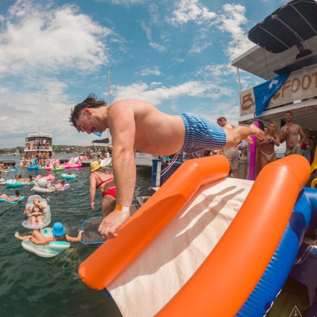 A man in blue shorts is diving off an inflatable slide into the water. People are swimming and lounging on floats in a sunny, outdoor marina setting.