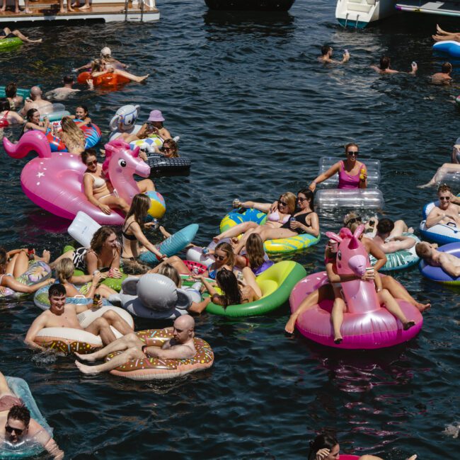 A large group of people floating in a lake on various inflatable rafts, including unicorns, donuts, and flamingos. The crowd is lively and enjoying the sunny day. Boats are visible in the background.
