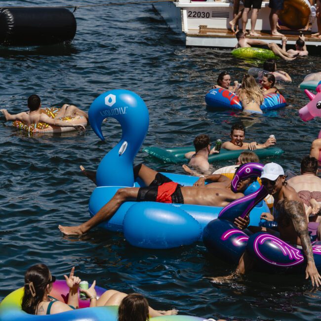 People are enjoying a sunny day on the water, relaxing on colorful inflatable floats. Boats are docked nearby with more people socializing on deck. The atmosphere is lively and festive.