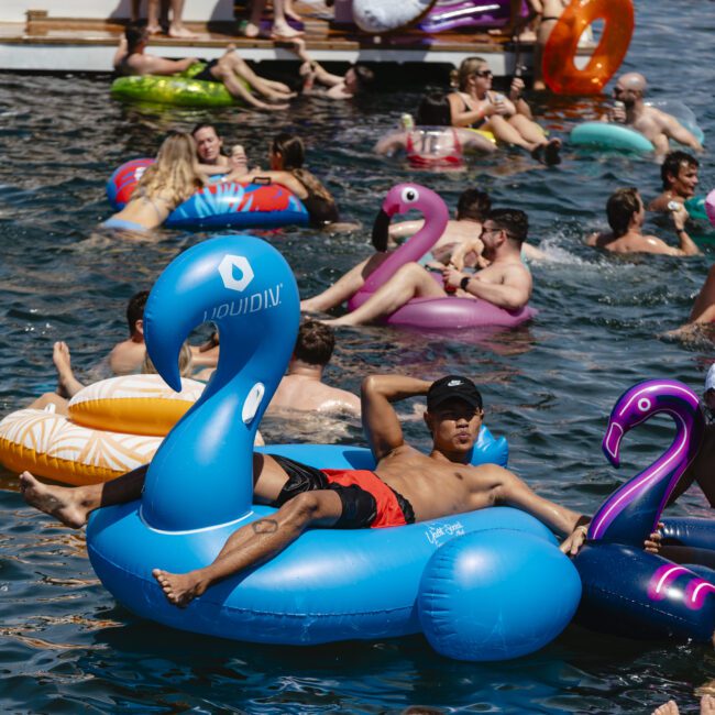 A group of people relax on colorful inflatable floats, including swans, flamingos, and a unicorn, in a body of water. Some are sunbathing, while others chat and enjoy the summer atmosphere.