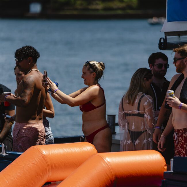 A group of people enjoying a sunny day on a boat by a lake. One person in a red bikini appears to be taking a selfie with a man in swim shorts. There's an inflatable slide in the foreground, and lush greenery is visible in the background.