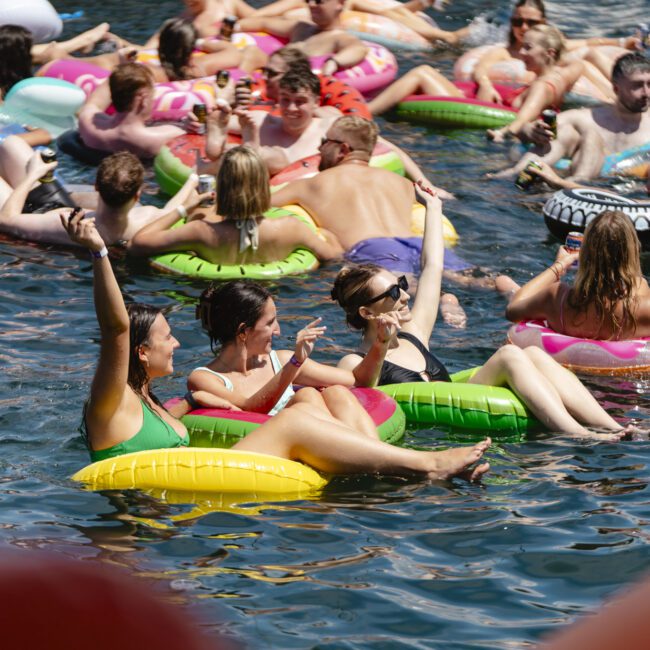 A crowded scene of people relaxing on colorful inflatable rings in a sunny body of water. The group appears to be enjoying the warm weather, with many holding drinks. A woman in the foreground raises her arm joyfully, while others chat and laugh.