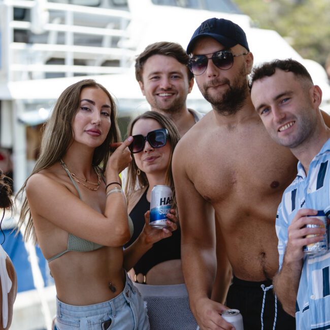 A group of five people smiling and posing together on a boat. Three men and two women are casually dressed, holding canned drinks. A boat's railing and part of the water are visible in the background.