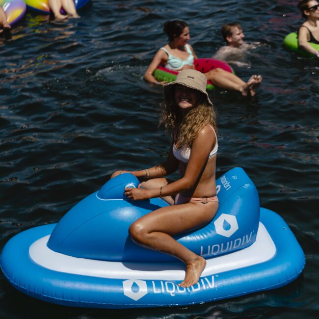 A person in a bikini and sun hat sits on a blue inflatable jet ski in a body of water. Around them, others are enjoying the water on various inflatables. The scene is lively and suggests a summer gathering or pool party.