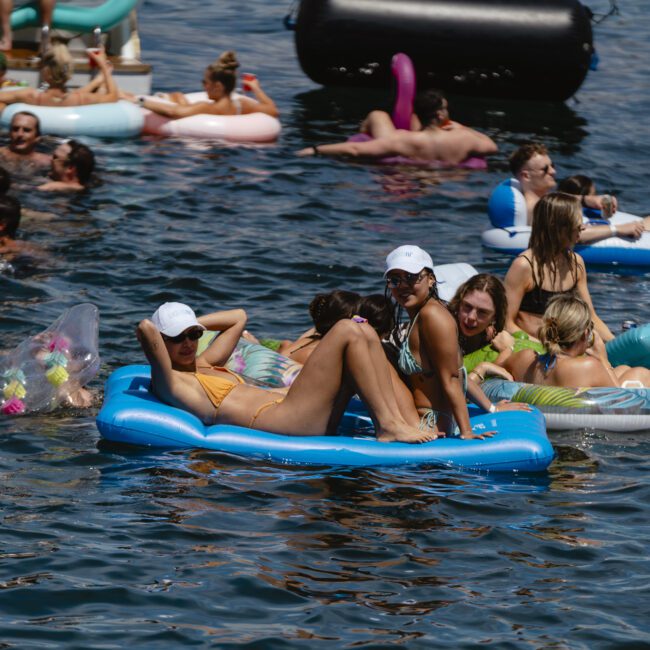 People relaxing on various inflatable floats in a crowded body of water, enjoying a sunny day. Some are wearing swimsuits and sunglasses, with inflatable flamingos and other pool toys visible in the background.