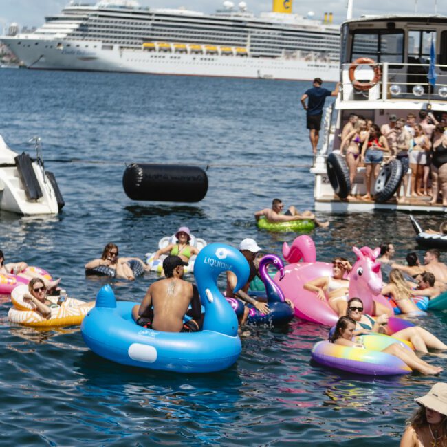 People are enjoying a sunny day on inflatable floaties shaped like animals in the water, near a boat. In the background, a large cruise ship is seen against a partly cloudy sky.